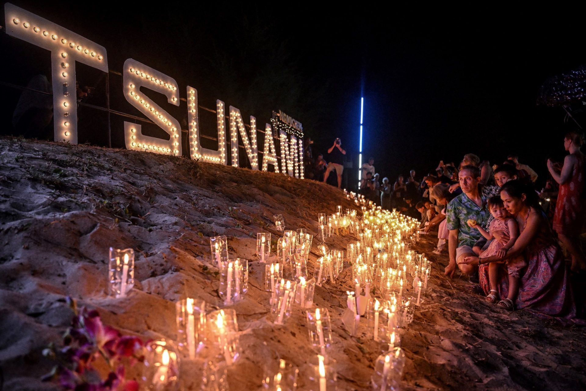 Night time - People placing candles on the sand in front of a lit Tsunami sign