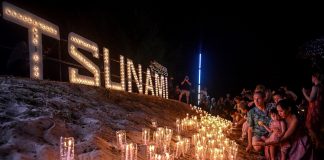 Night time - People placing candles on the sand in front of a lit Tsunami sign