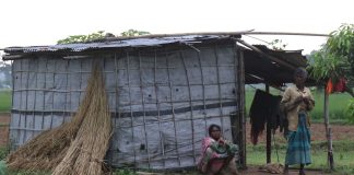 Two Indigenous persons sitting and standing next to a dilapidated looking hut in rural Bangladesh